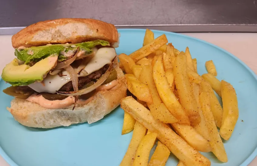 A Hamburguesas Emilia burger topped with sliced avocado, melted white cheese, grilled onions, and chipotle mayo on a toasted bun, with seasoned fries.