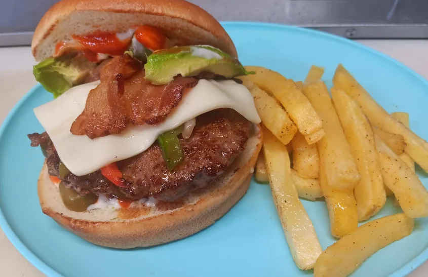 A burger layered with crispy bacon, white cheese, and avocado on a toasted bun, served with seasoned fries.