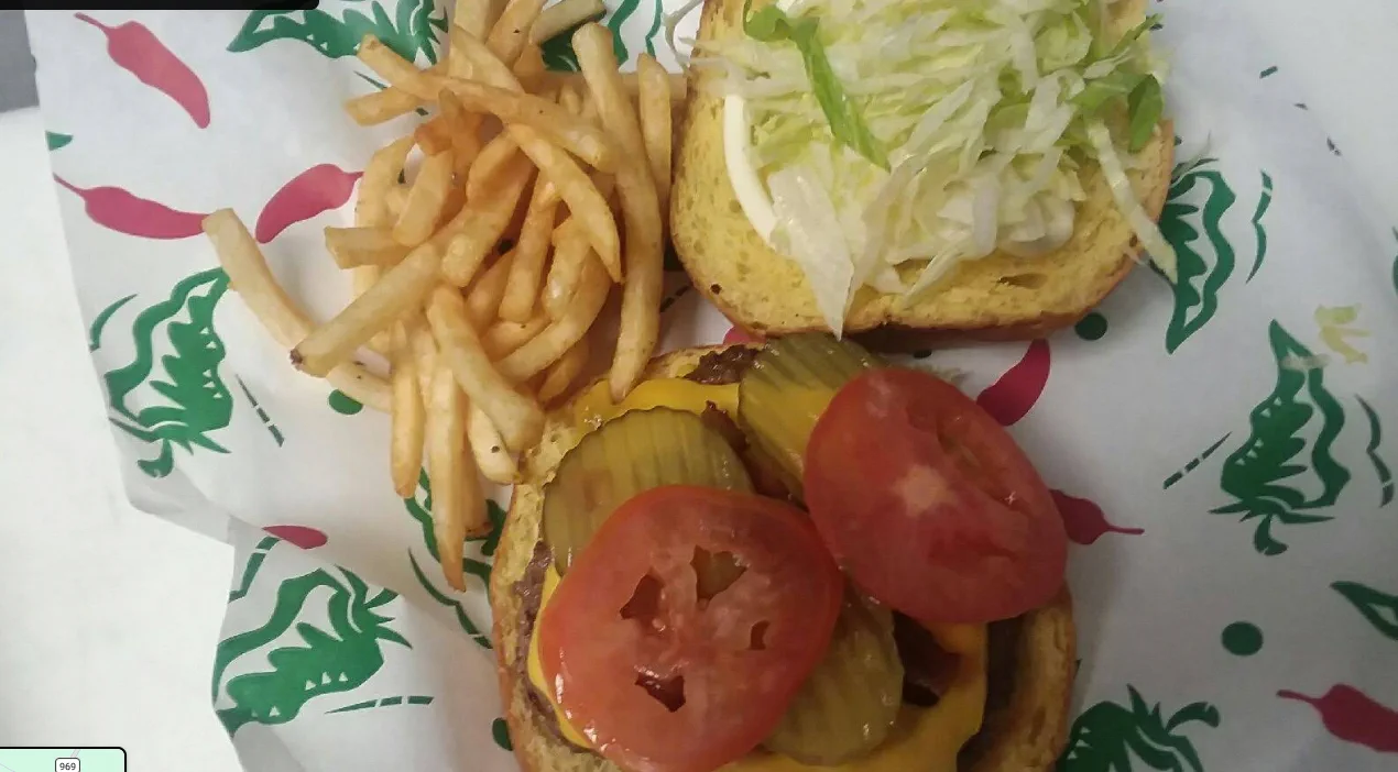An open-faced cheeseburger on chile-pepper printed paper, stacked with melted cheese, pickles, fresh tomato, and shredded lettuce, with shoestring fries alongside.