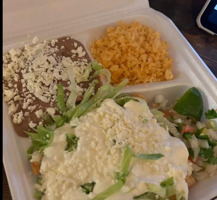 A plate of chicken flautas topped with lettuce, crema, and crumbled cheese, with refried beans, Mexican rice, pico de gallo, and a lime wedge.