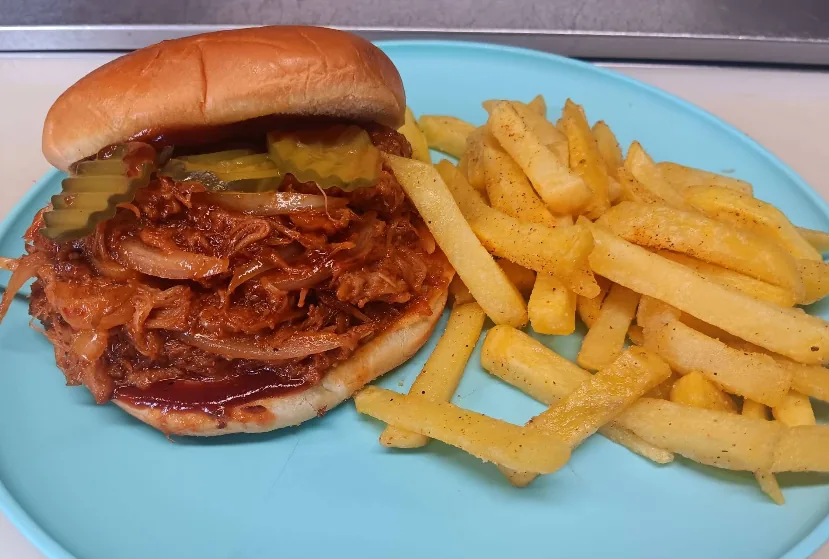 A pulled pork sandwich glazed in BBQ sauce with pickles and onions, served with seasoned fries.