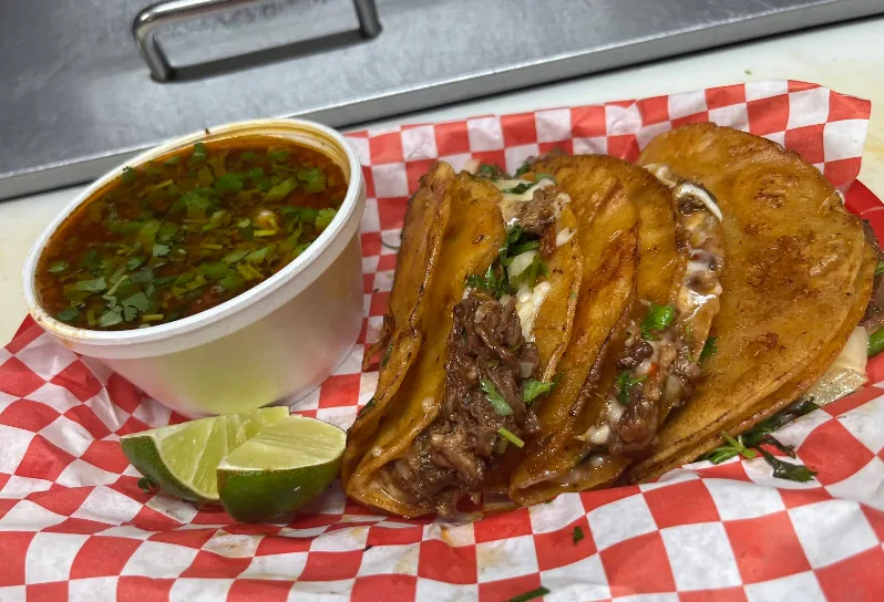 Quesabirria tacos with crispy cheese edges next to a cup of consomé topped with cilantro and lime.