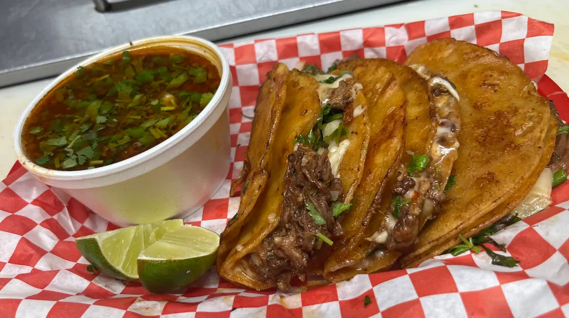 Three griddle-crisped quesabirria tacos with melted cheese and tender birria beef on red-and-white checkered paper, with a cup of rich consomé and lime wedges.