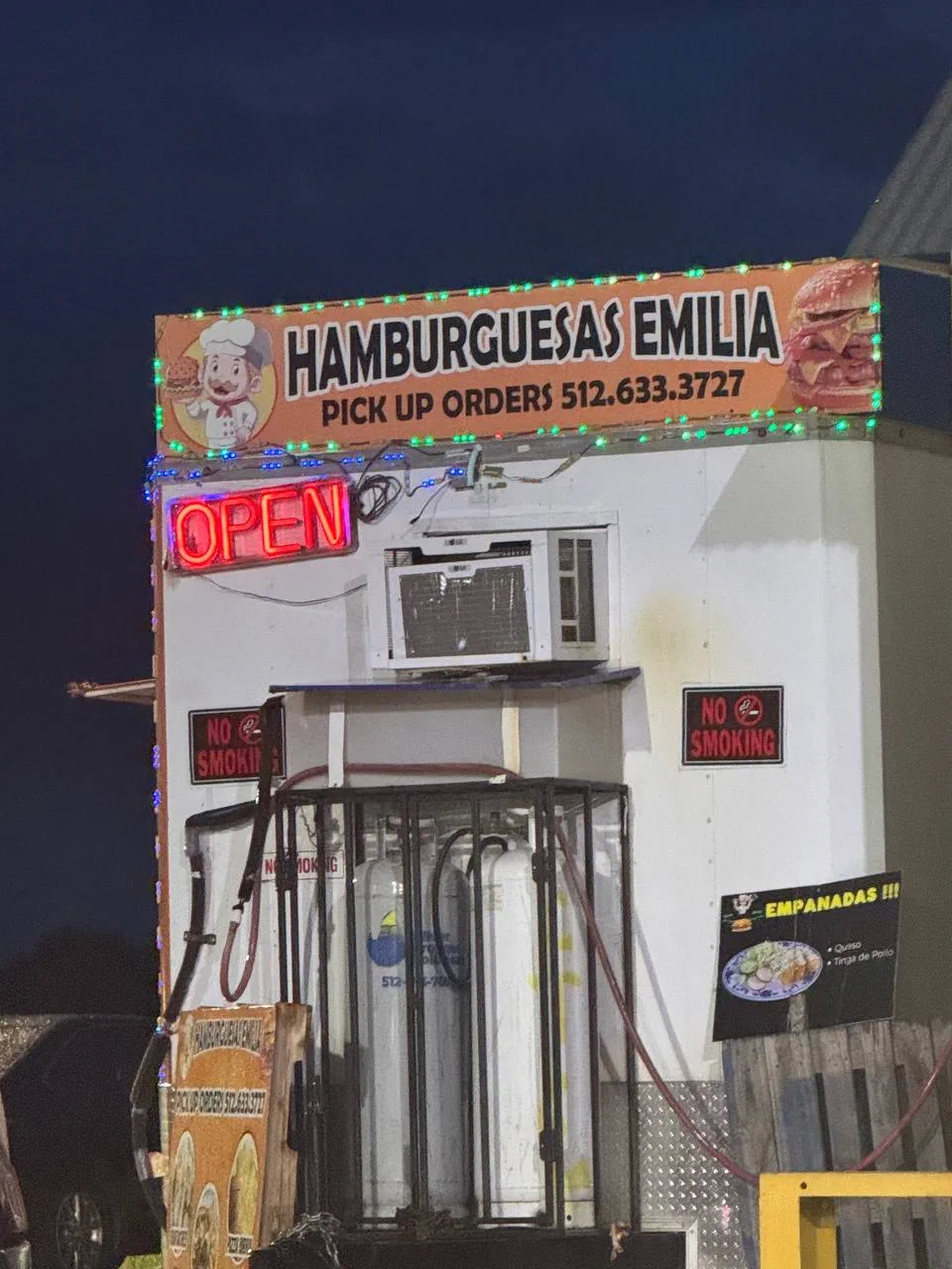 The lit Hamburguesas Emilia sign at night with a neon OPEN sign and 'Pick up orders 512.633.3727', on the food truck on FM 969.