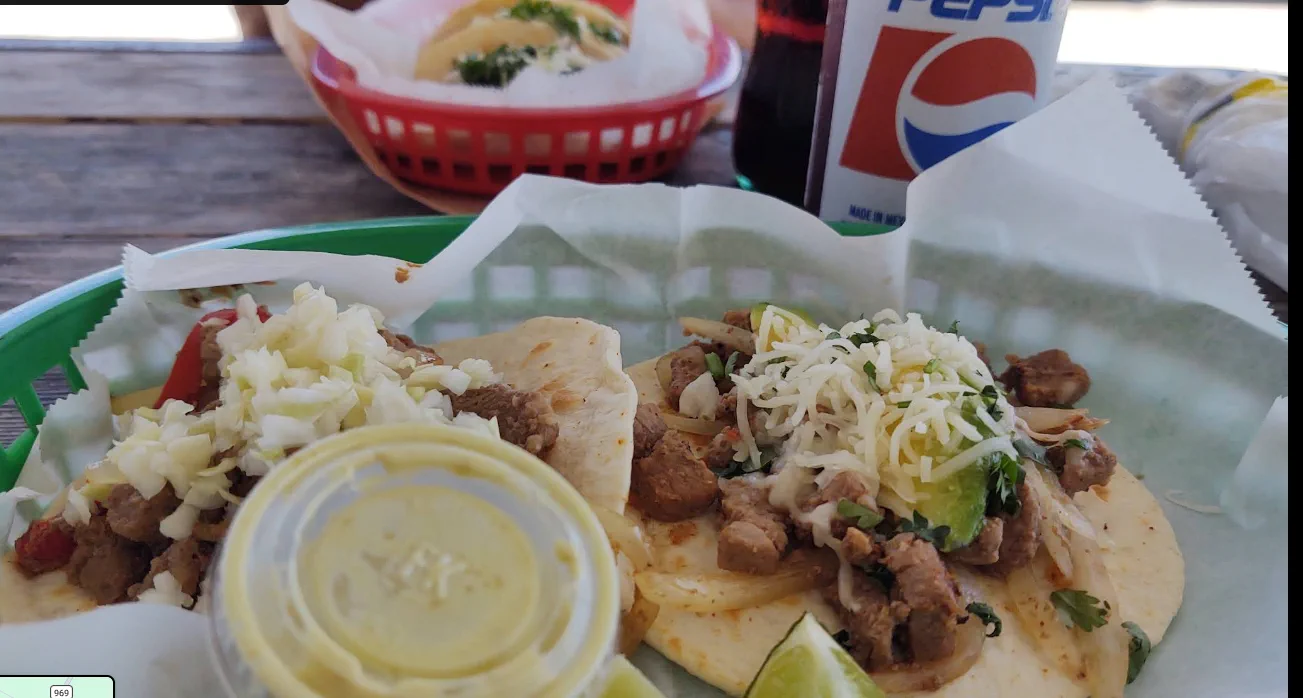 Two street tacos in a green basket, topped with grilled steak, onion, cilantro, cheese, and avocado, with lime and a cup of salsa.